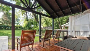 A covered outdoor deck with wooden chairs and a table, surrounded by greenery and a house in the background.