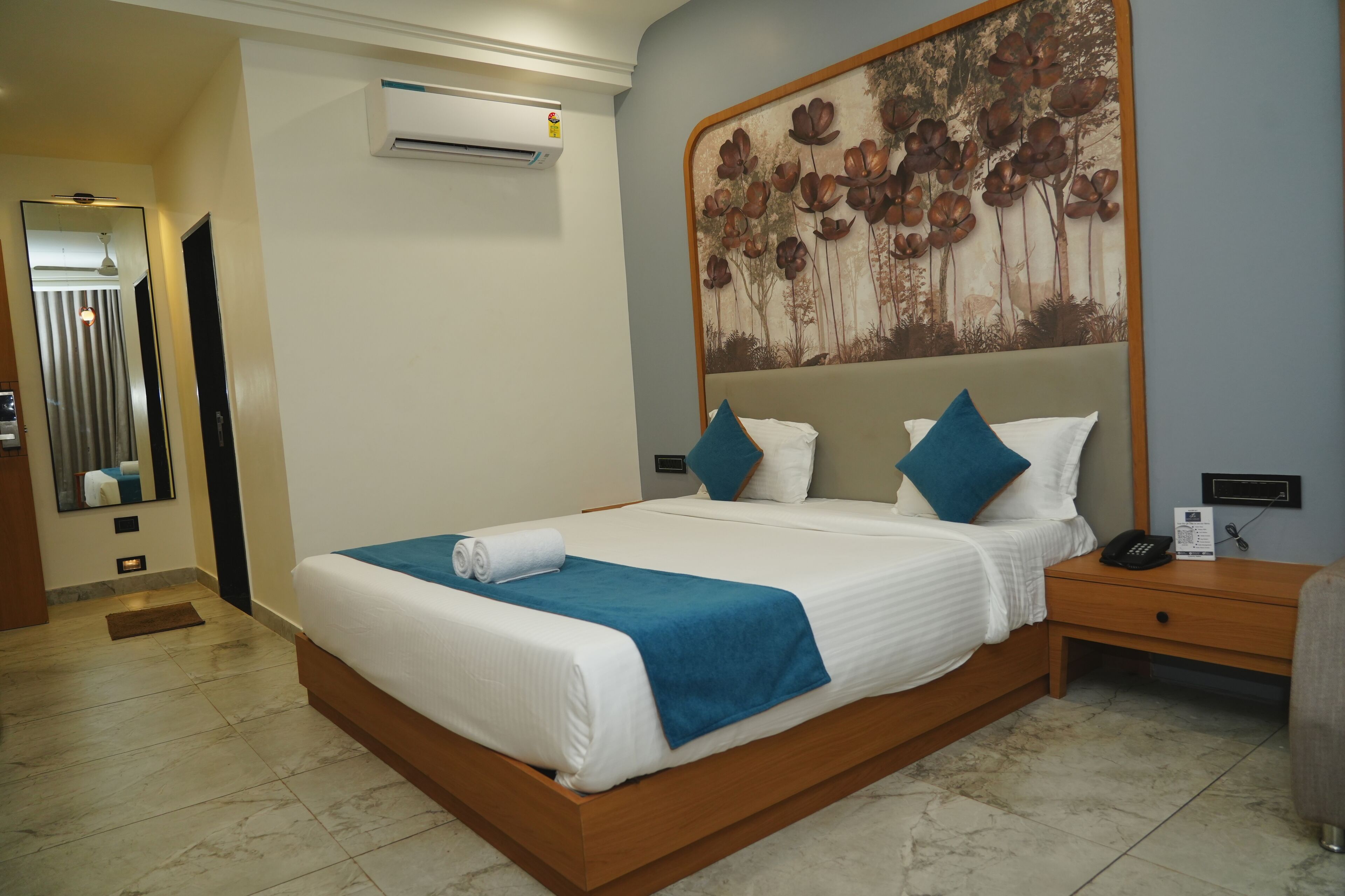 A hotel room with a large bed, a nightstand, an air conditioning unit, and a wall-mounted artwork.