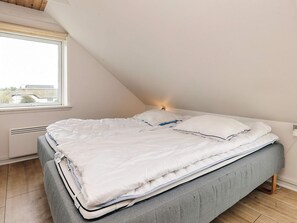 A bedroom with a sloped ceiling, a bed with white bedding, a grey upholstered bench, and a window with a view of buildings.