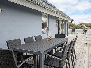 Een overdekte eethoek met een tafel en stoelen, een vaas met bloemen, en een uitzicht op een huis op de achtergrond.