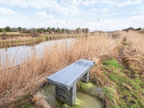 A stone bench by a calm water body, surrounded by tall grasses and a clear sky.