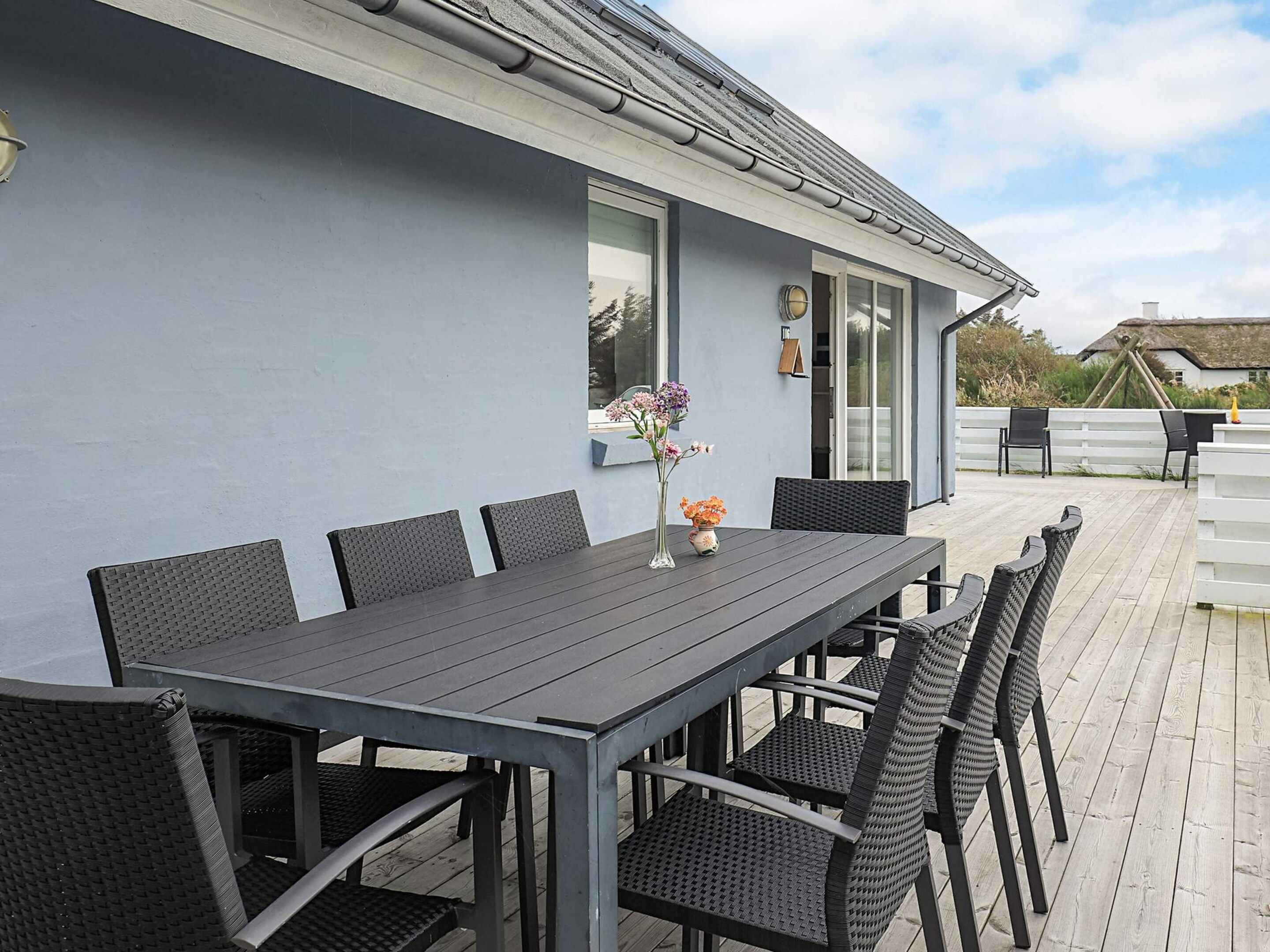 A modern outdoor dining area with a grey table and black wicker chairs, a white fence, and a clear sky.