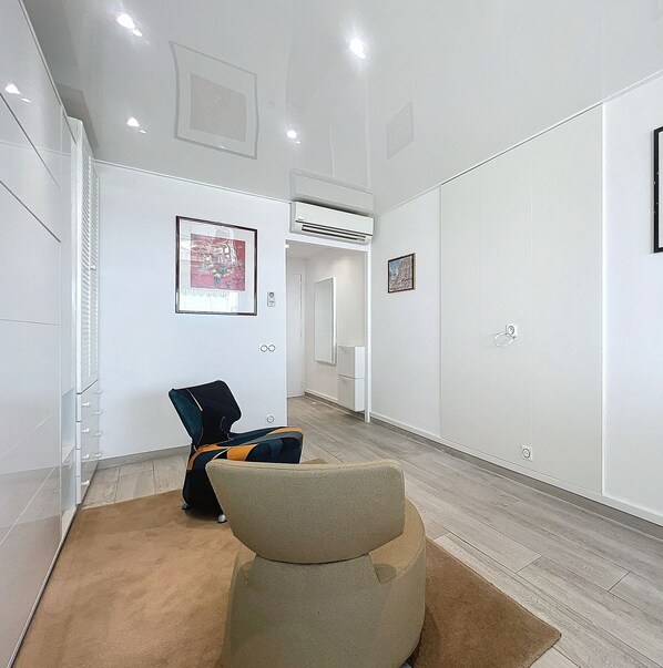 Un couloir avec un fauteuil beige, un fauteuil noir et orange, et un tapis marron. Des tableaux encadrés ornent les murs et un appareil de climatisation est fixé au plafond.