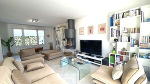 A modern living room with a beige sofa, a glass coffee table, a flat-screen TV, a fireplace, and a bookshelf filled with books. - Contemporary villa 100m from the beach with garden and WIFI (Trégastel)