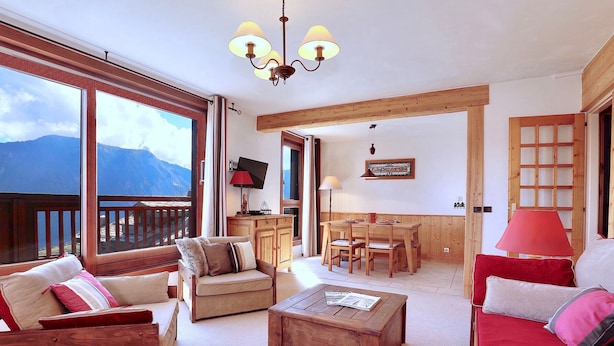 Ein geräumiges Wohnzimmer mit einem Holztisch, einem roten Sofa und einem beigefarbenen Sofa mit Kissen. Es gibt einen Holzessttisch mit Stühlen, einen an der Wand montierten Fernseher und ein großes Fenster mit Blick auf die Berge.