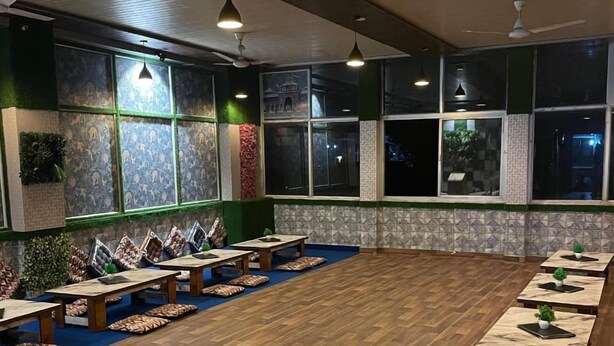 A spacious indoor area with a long row of wooden tables and benches, large windows with patterned curtains, and ceiling fans. - Namoh Stay (Rishikesh)