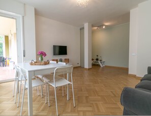 A modern living room with a white dining table, white chairs, a flat-screen TV mounted on the wall, and a grey sofa. - Turati Family Apartment (Lecco)