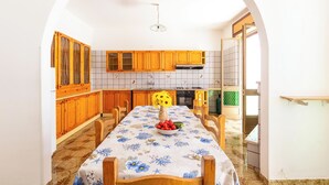 Une cuisine avec des meubles en bois, un dosseret carrelé et une table à manger recouverte d’une nappe à motifs floraux et agrémentée d’un saladier de fruits.