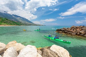 Kayakers in green kayaks on a clear turquoise sea with rocky shores and mountains in the background.