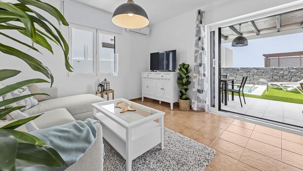 Una sala moderna con sofá, mesa de centro, televisor y vista a un patio con mobiliario exterior.