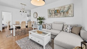 Una sala moderna con un sofá blanco, una mesa de centro de vidrio y un comedor con una mesa y sillas blancas.