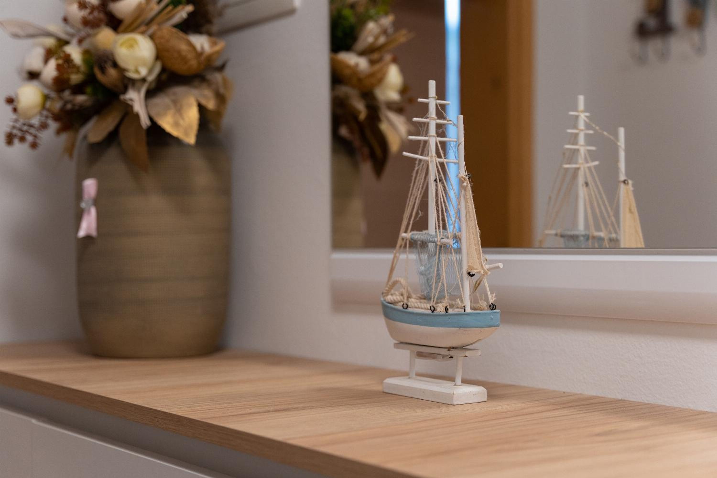 A model ship on a wooden shelf with a vase of dried flowers in the background.