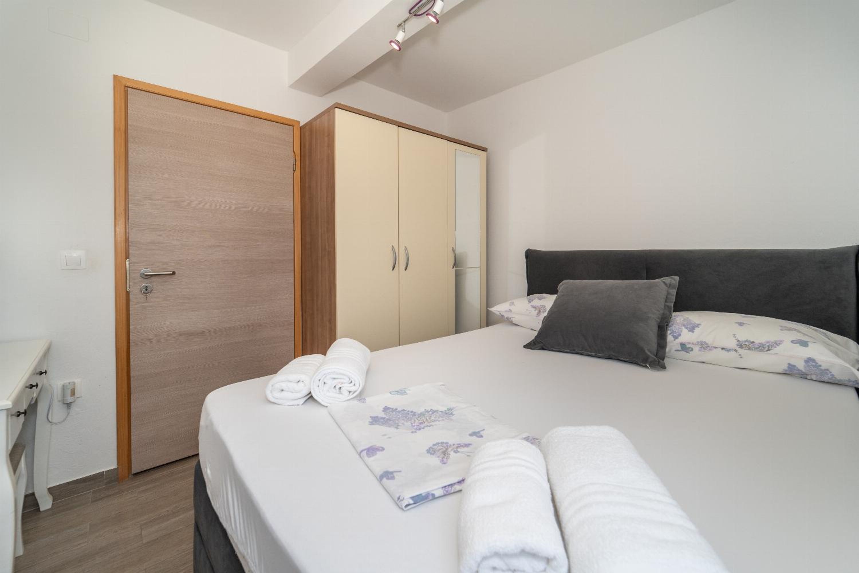 A neatly made bed with a floral bedspread, a grey headboard, and two pillows. There is a wooden wardrobe with a mirror and a white bedside table with a lamp.