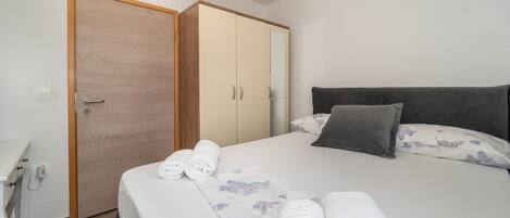A neatly made bed with a floral bedspread, a grey headboard, and two pillows. There is a wooden wardrobe with a mirror and a white bedside table with a lamp.