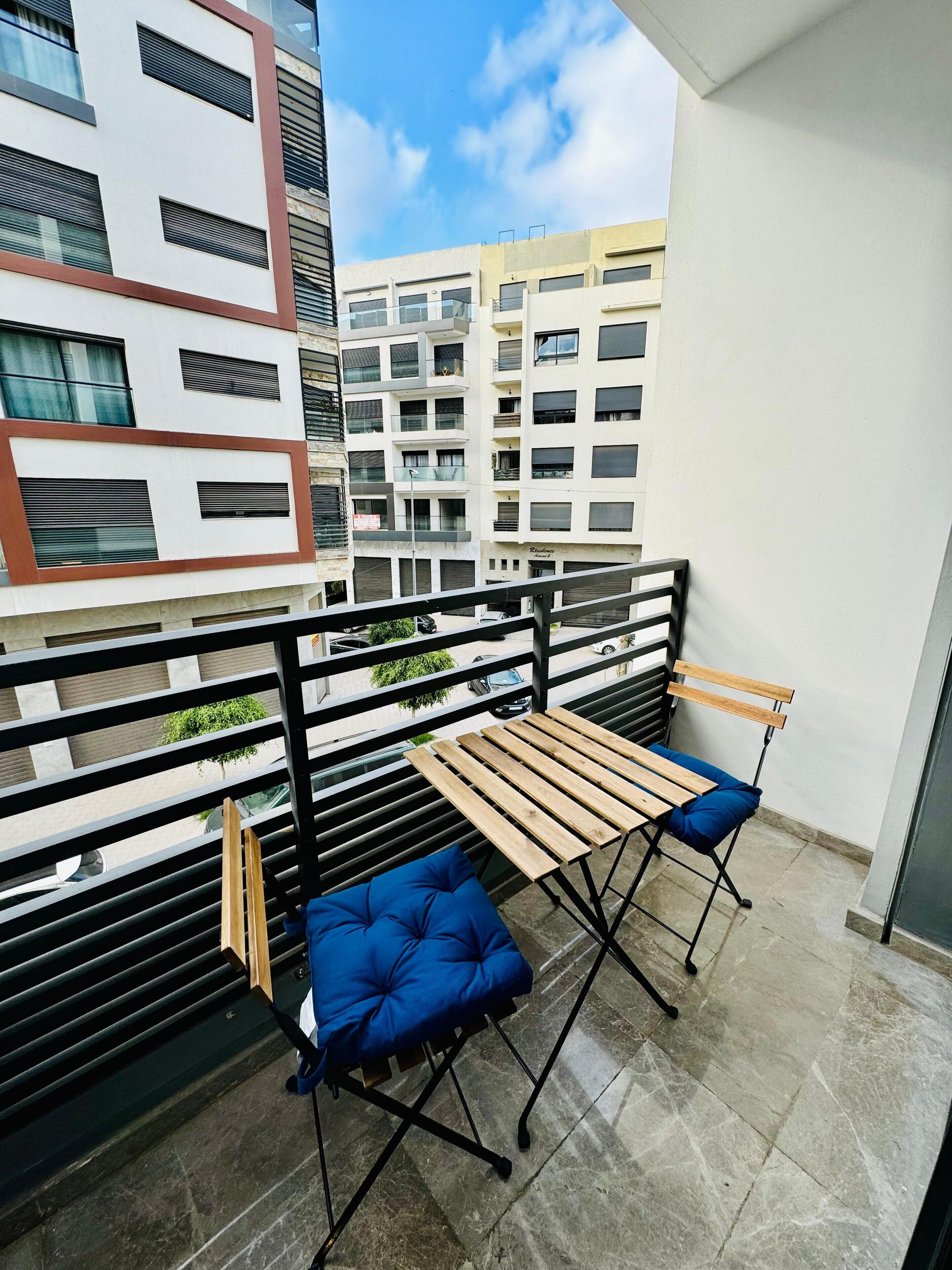 A balcony with a wooden table and blue cushioned chairs, overlooking a cityscape with modern buildings.