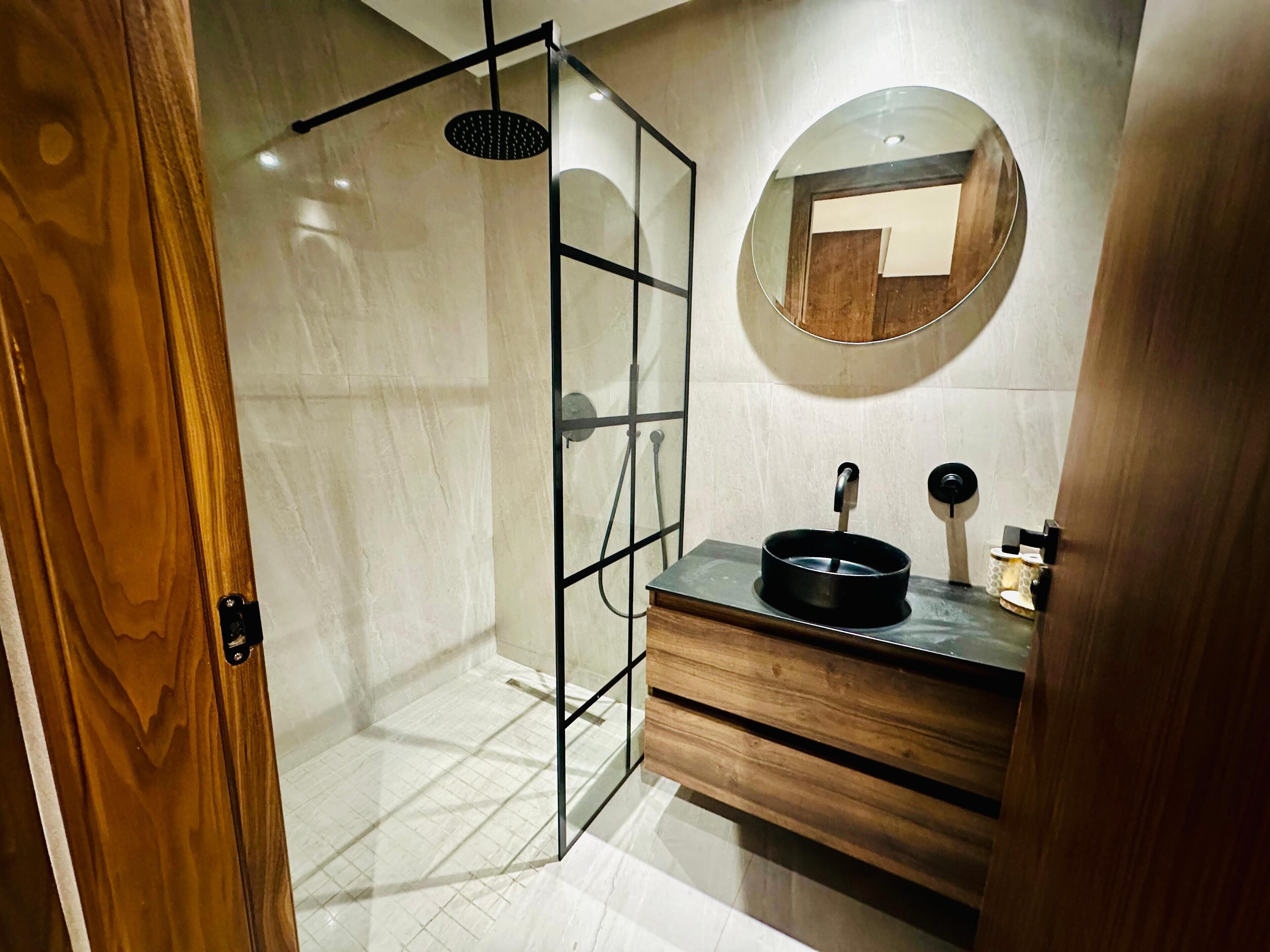 Un bagno moderno con box doccia in vetro, un mobile lavabo in legno e uno specchio rotondo.