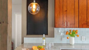 A kitchen with wooden cabinets, a marble countertop, a hanging light fixture, a wine bottle, a vase with tulips, and a fruit platter.