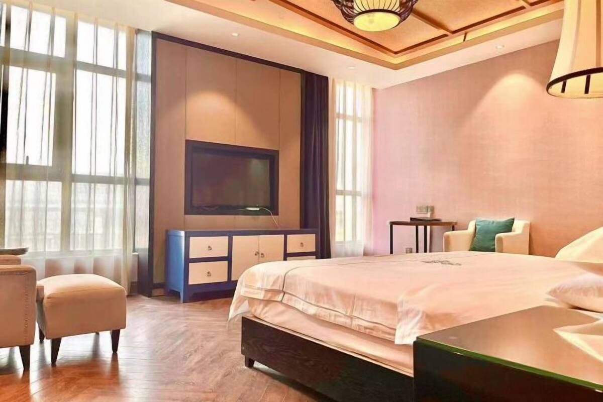 A modern hotel room with a large bed, a flat-screen TV mounted on the wall, a small side table, and a seating area with a chair and ottoman.