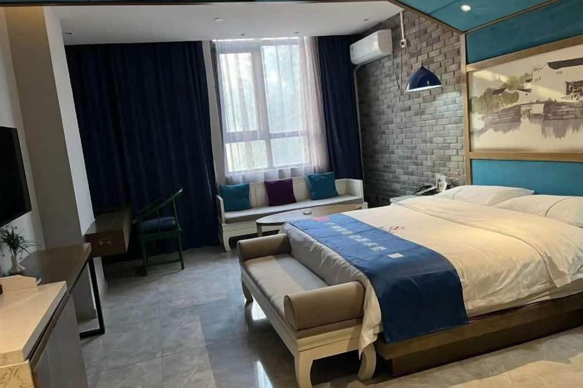 A hotel room with a bed, a desk, a chair, a bench, a window with curtains, and a wall-mounted air conditioner.