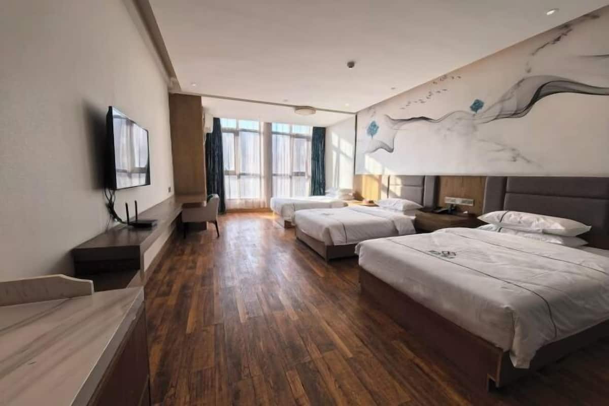 A hotel room with a wooden floor, three beds, a TV on the wall, and a large window with curtains.
