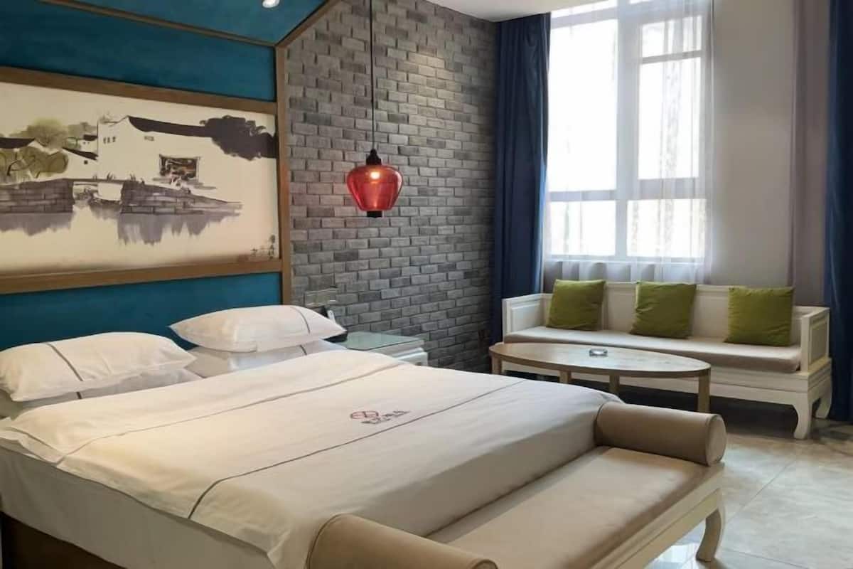 A hotel room with a bed, a sofa, a small table, a hanging light, and a wall with a mural.