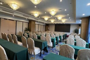 Une salle de conférence avec des rangées de tables et de chaises, un grand écran et des lustres.