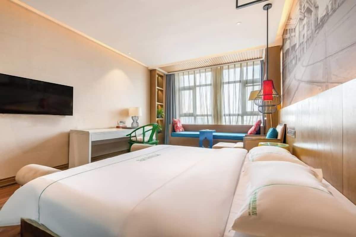 A hotel room with a large bed, a desk, a chair, a TV mounted on the wall, and a window with curtains.