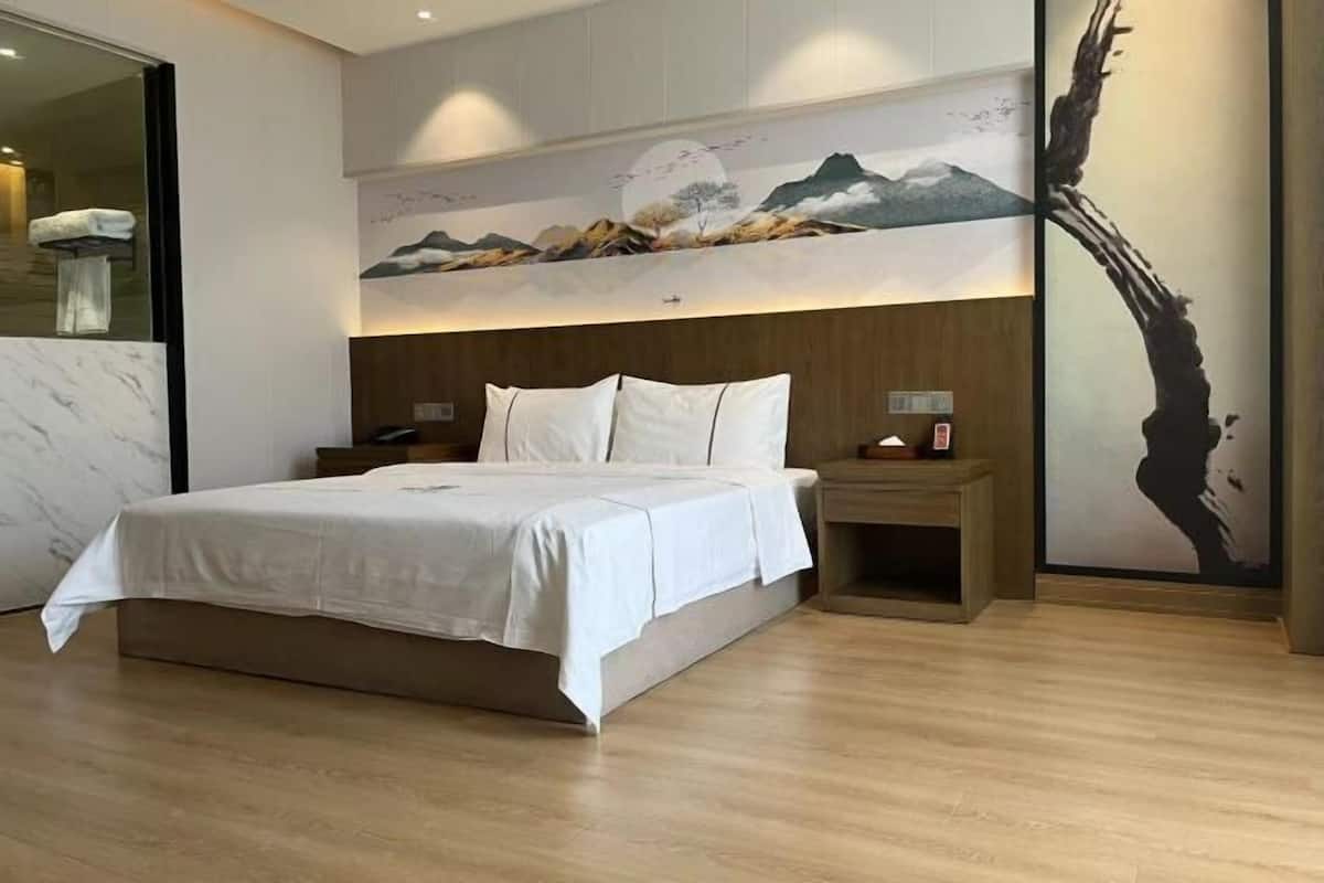 A modern hotel room with a large bed, wooden headboard, and a painting of mountains on the wall.