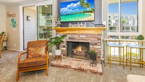 A living room with a fireplace, a TV mounted above it, and a view of a beach through large windows. - NCV C4 Beautiful beachfront cottage, all new furnishings w/ Stunning Ocean views (Oceanside)