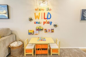 A child's play area with a wall sign that reads 'where the WILD one play' and a table with educational toys and books.