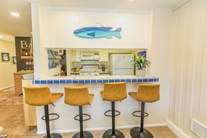 Uma área de bar com decoração de peixe azul, micro-ondas e geladeira. Há quatro banquetas e uma planta em vaso.