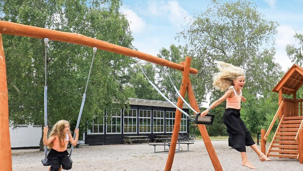 A playground with swings and a slide, surrounded by trees and a building with windows.