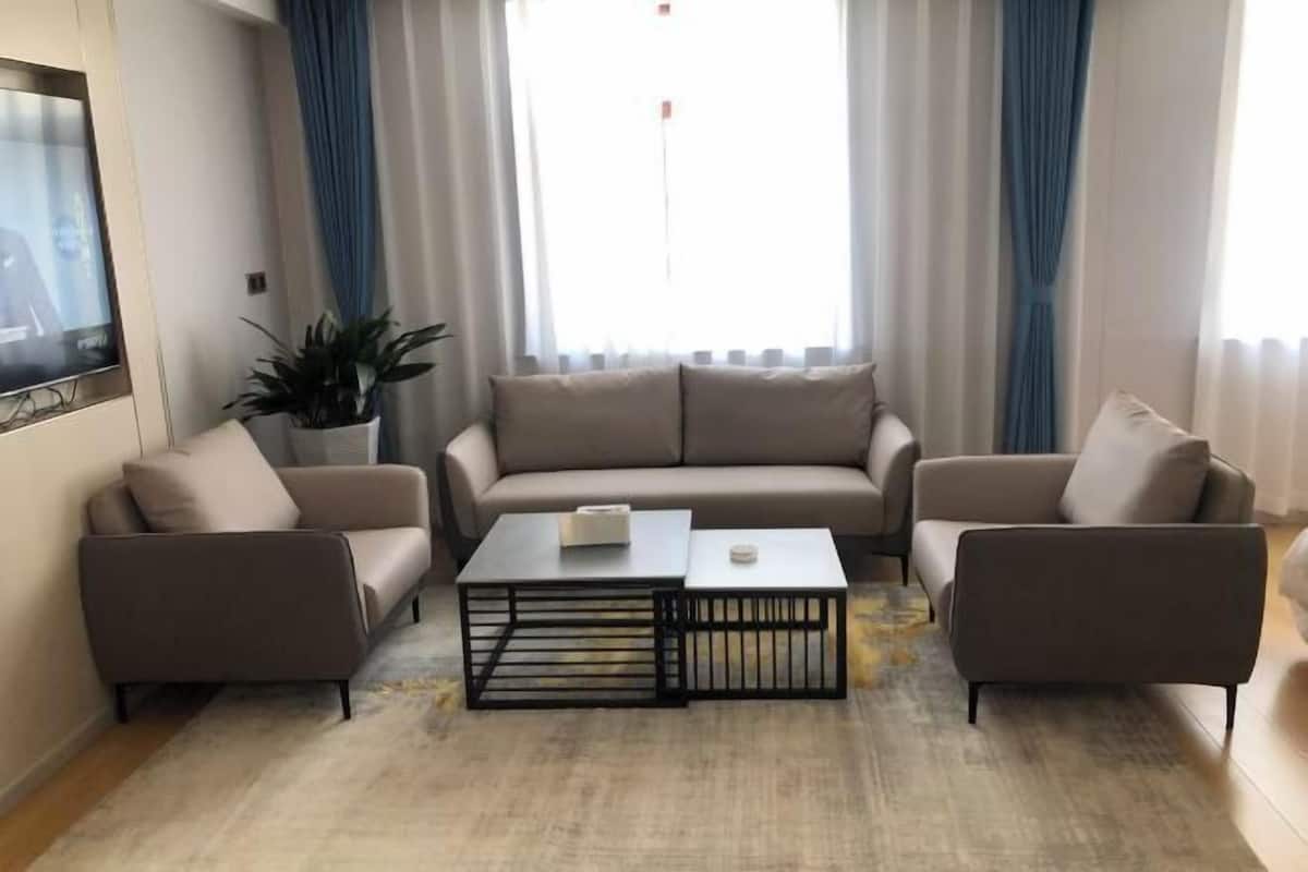 Ένα σαλόνι με έναν καναπέ, δύο πολυθρόνες, ένα τραπεζάκι σαλονιού, μια τηλεόραση και ένα παράθυρο με κουρτίνες.
