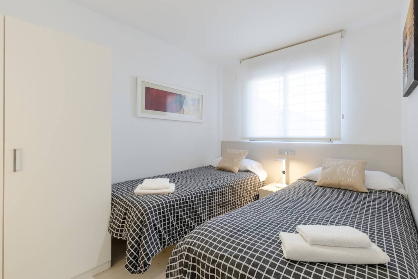 A room with two beds, each with a checkered bedspread and a pillow. There is a window with blinds, a bedside table with a lamp, and a framed picture on the wall.