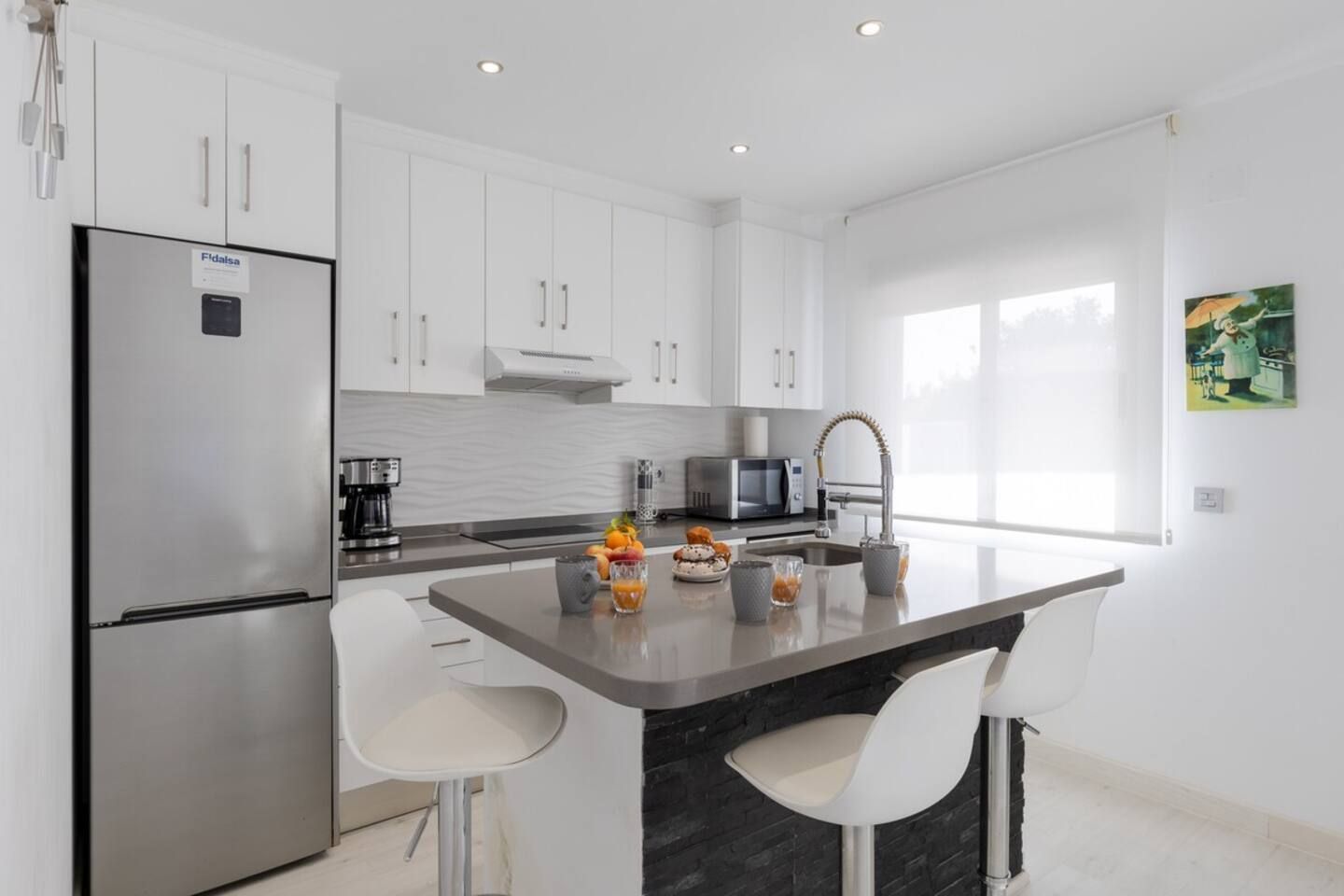 Een moderne keuken met witte kasten, een roestvrijstalen koelkast, een magnetron en een spoelbak met een moderne kraan. Er staan twee witte barkrukken bij een klein aanrecht met fruit en drankjes.
