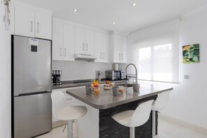 Cocina moderna con armarios blancos, un refrigerador de acero inoxidable, un microondas y un lavamanos con grifo moderno. Hay dos taburetes de bar blancos y una pequeña isla con fruta y bebidas.