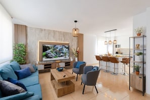 A modern living room with a flat-screen TV, a wooden coffee table, and a sofa. There is a dining area with bar stools and a kitchen visible in the background.