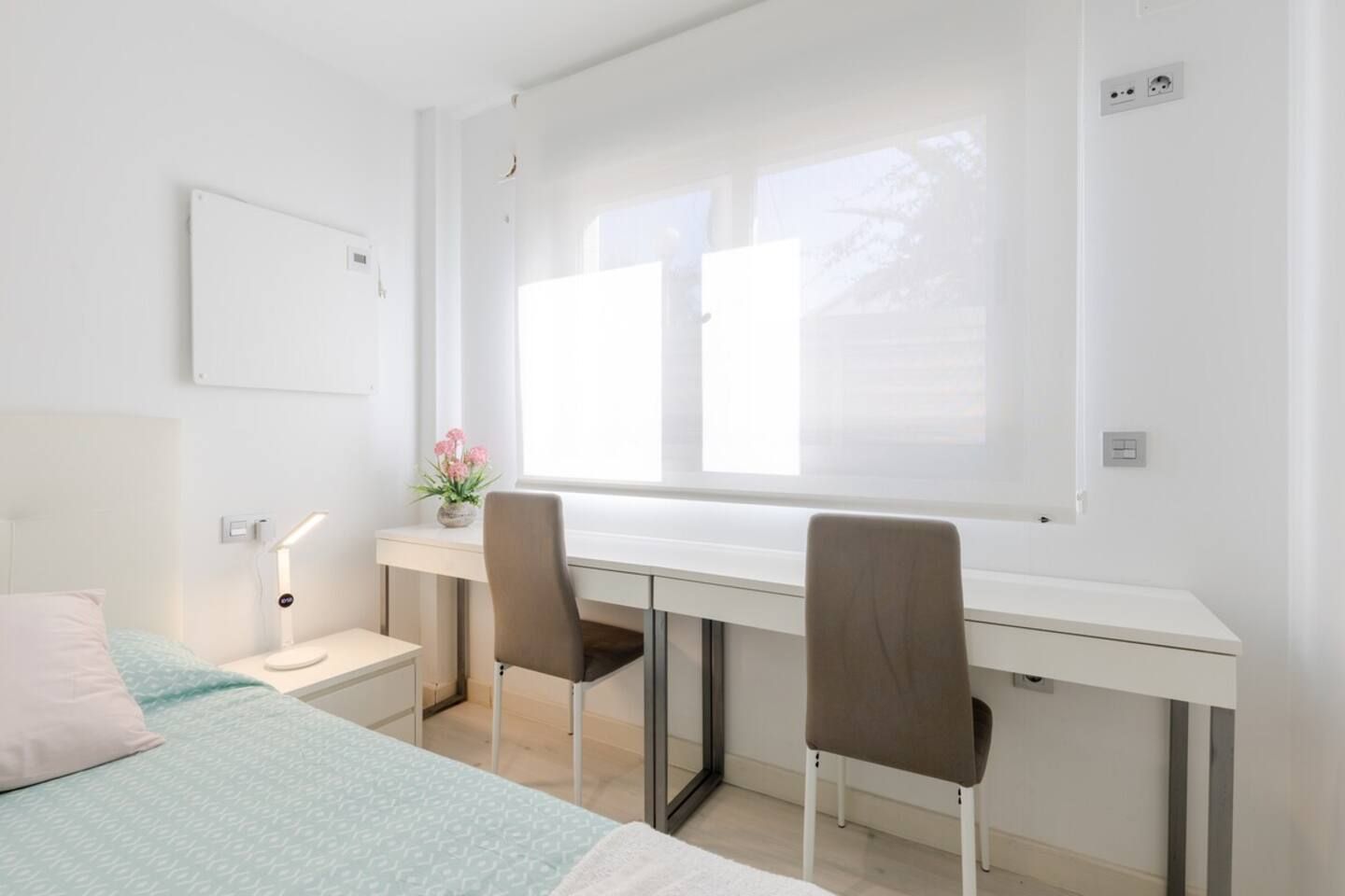 Un dormitorio moderno y bien iluminado con cama, escritorio, dos sillas y ventana que proporciona luz natural.