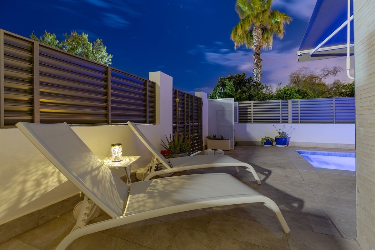 Een modern terras buiten met twee loungestoelen, een bijzettafeltje met een lamp, een zwembad en een palmboom.