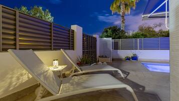 Een modern terras buiten met twee loungestoelen, een bijzettafeltje met een lamp, een zwembad en een palmboom.