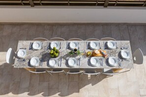 A rectangular outdoor dining table with white chairs, set with plates, cutlery, and a centerpiece of green vegetables.