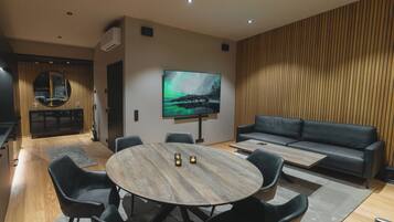 Een moderne woonkamer met een ronde houten tafel, een zwarte leren bank en een flatscreen televisie aan de muur.