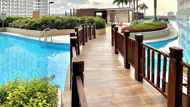 A pool area with a wooden deck, surrounded by greenery and a view of buildings.