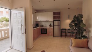 A modern kitchen with white cabinets, a built-in oven, and a refrigerator. There is a dining area with a table and chairs, and a large plant in the corner. - Chill Out Apartments (Chania)