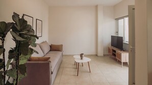 A living room with a sofa, a coffee table, a TV on a wooden stand, and large green plants. - Chill Out Apartments (Chania)