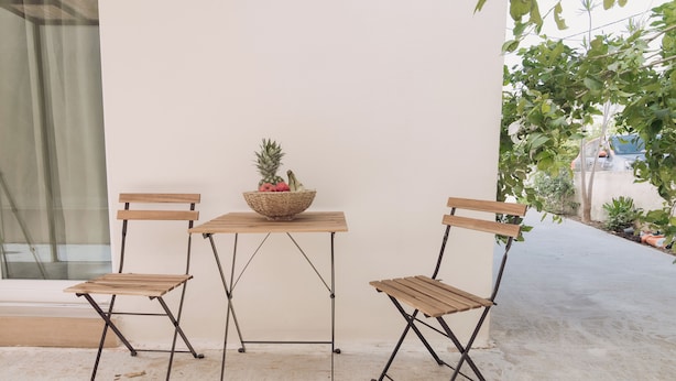 A small outdoor seating area with two wooden chairs and a table, a woven basket with fruits on the table, and a concrete floor. - Chill Out Apartments (Chania)