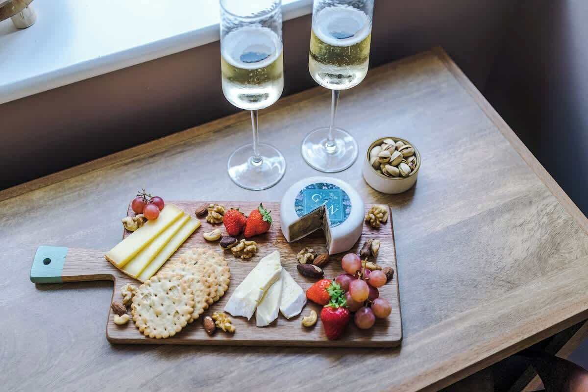 A wooden table with a cheese board, grapes, nuts, and two glasses of white wine.