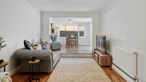 A modern living room with a grey sofa, a small side table, and a wooden cabinet. There is a dining area with a wooden table and chairs in the background. - Knights Rest - Minutes to the Port (Dover)