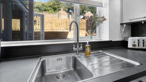 A modern kitchen with a stainless steel sink, a window with a view of a wooden fence and trees, and a toaster on the counter. - Knights Rest - Minutes to the Port (Dover)