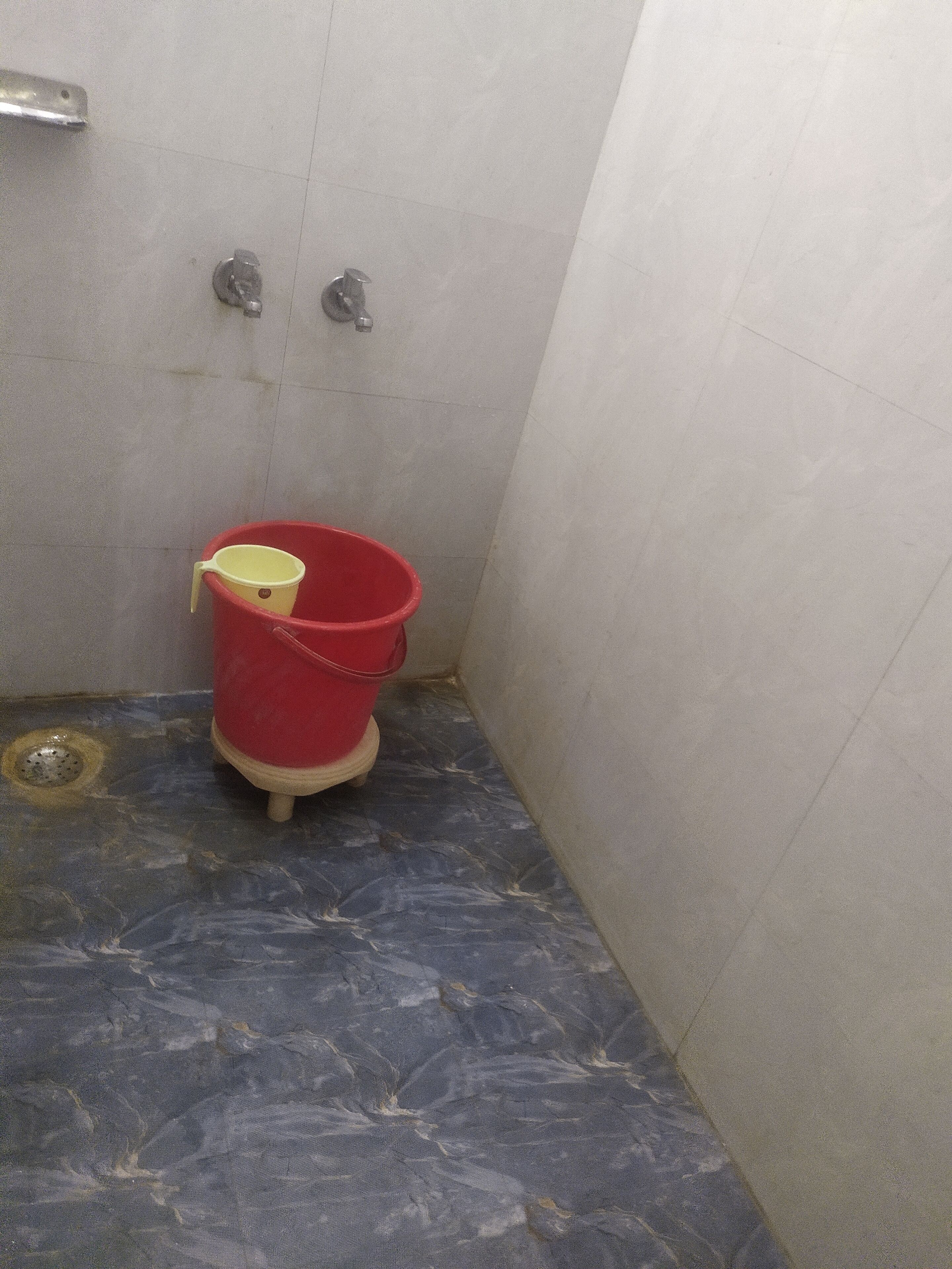 A small bathroom with a red bucket, a yellow cup, and a metal showerhead.